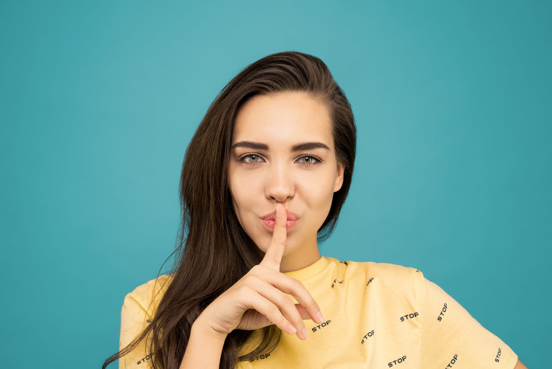 portrait photo of woman in yellow t shirt doing the shh sign while standing in front of blue background
